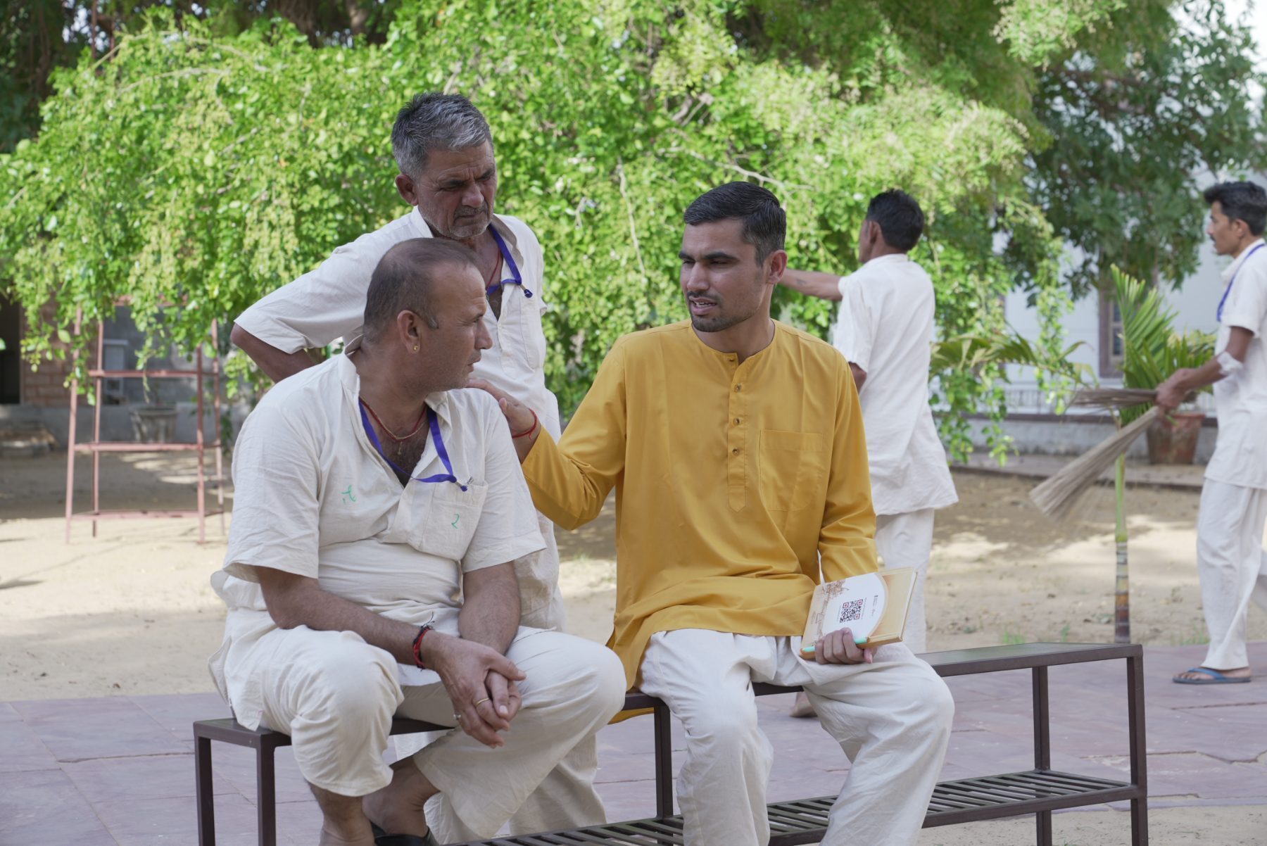 Two men sitting on a bench in a sunlit courtyard. One rests a hand on the other's shoulder and speaks quietly to him. A third stands close by, listening.