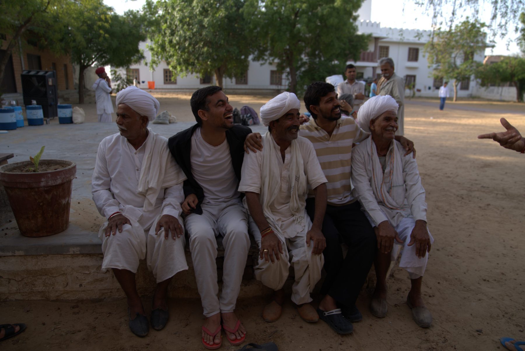 The next day in sunlight: elders in white turbans and members of the team sitting together on a low wall, arms around each other's shoulders, all of them laughing.