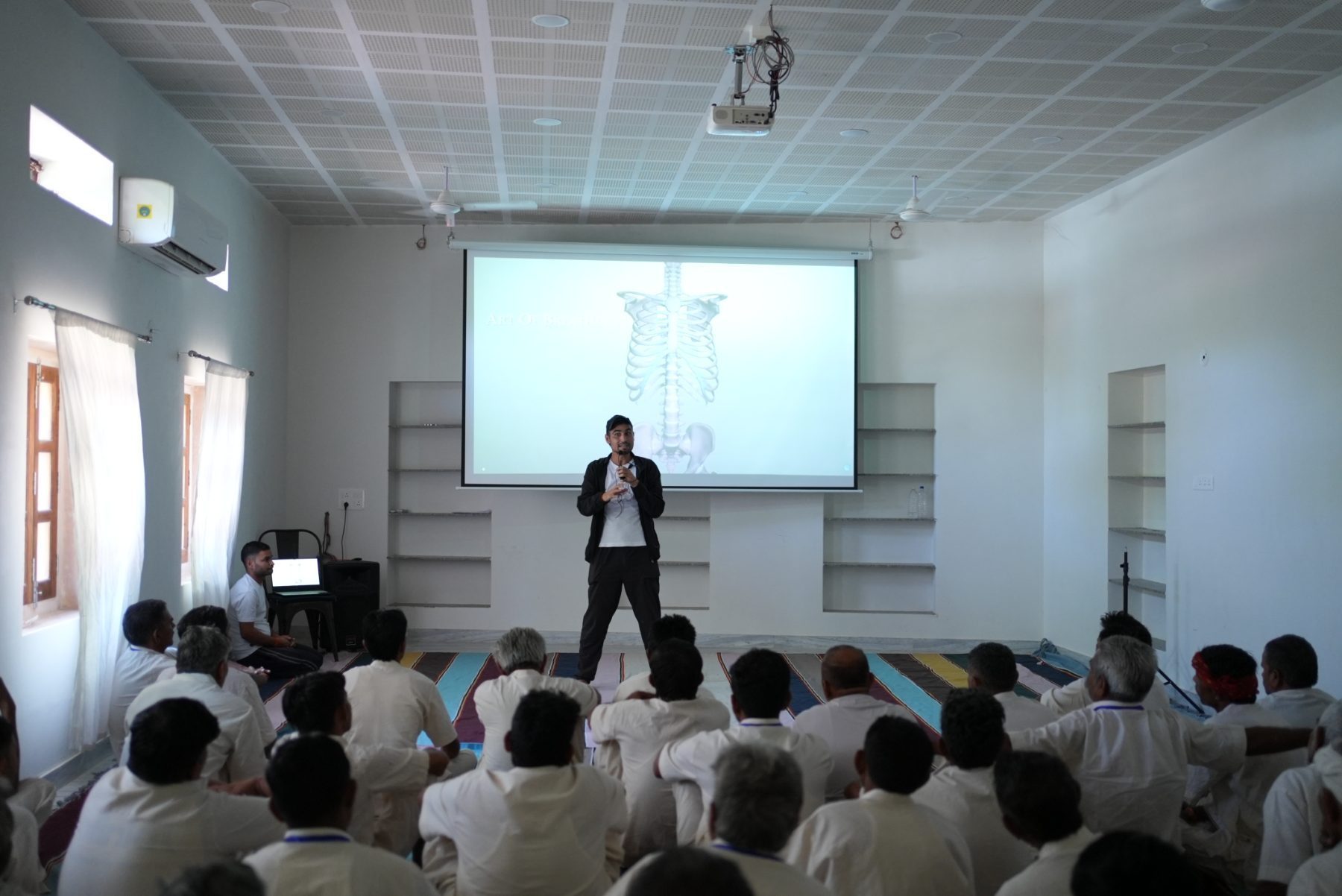 A teacher explaining a diagram of the ribcage projected on a screen, to men seated on mats.