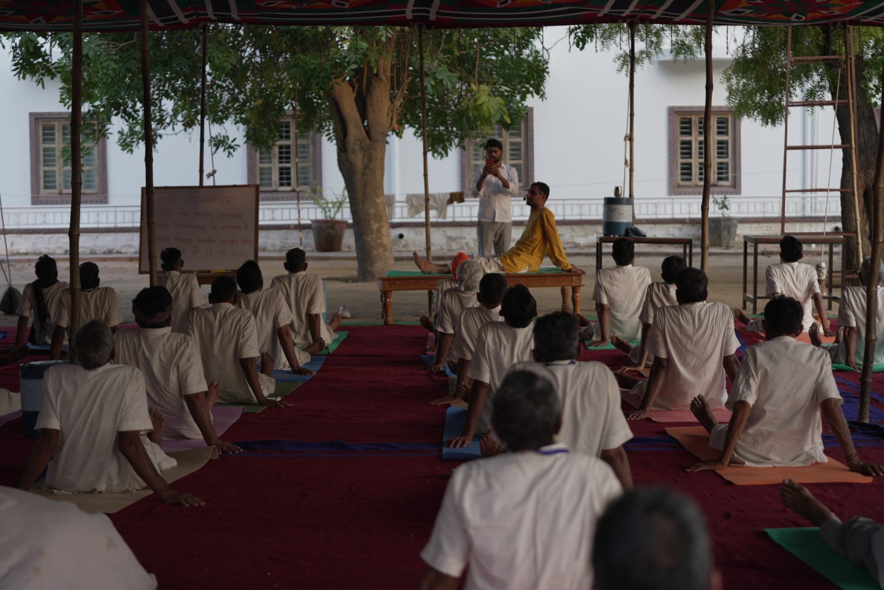 A class under a canopy in the dim early light, everyone seated with their backs to the camera, an instructor demonstrating on a low bench beside a whiteboard.