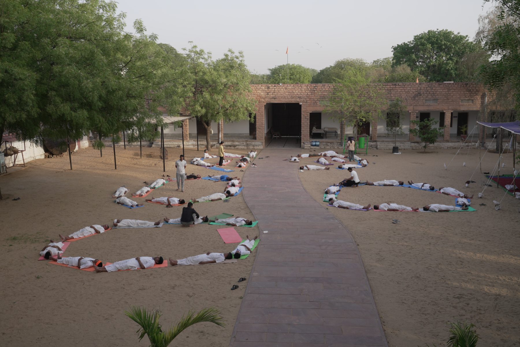Men lying in shavasana in a sand courtyard arranged in a wide arc, seen from above.