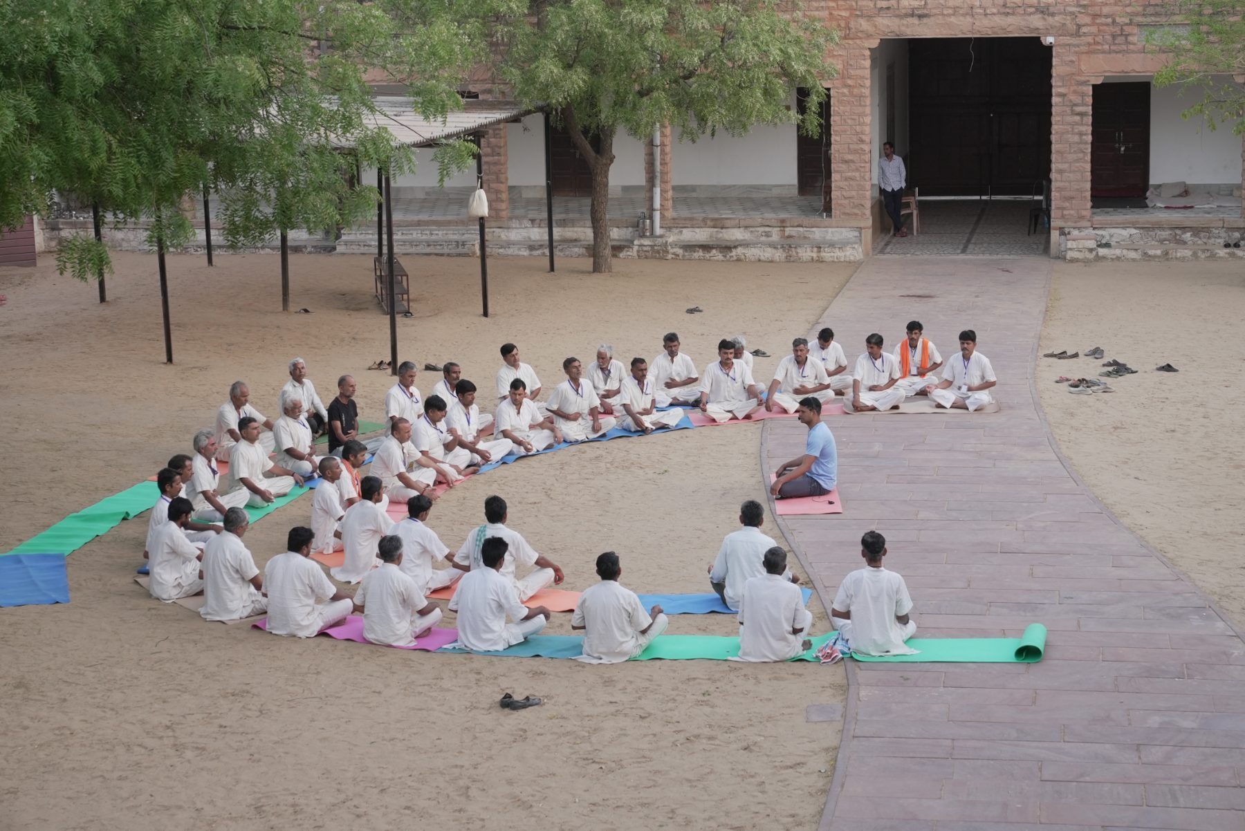 Men seated in rows on mats in a sand courtyard, facing a single instructor.