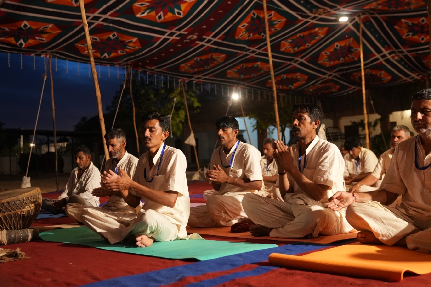 Men seated on the ground under a patterned canopy at night, hands joined, eyes closed, a drum at the edge of the frame.