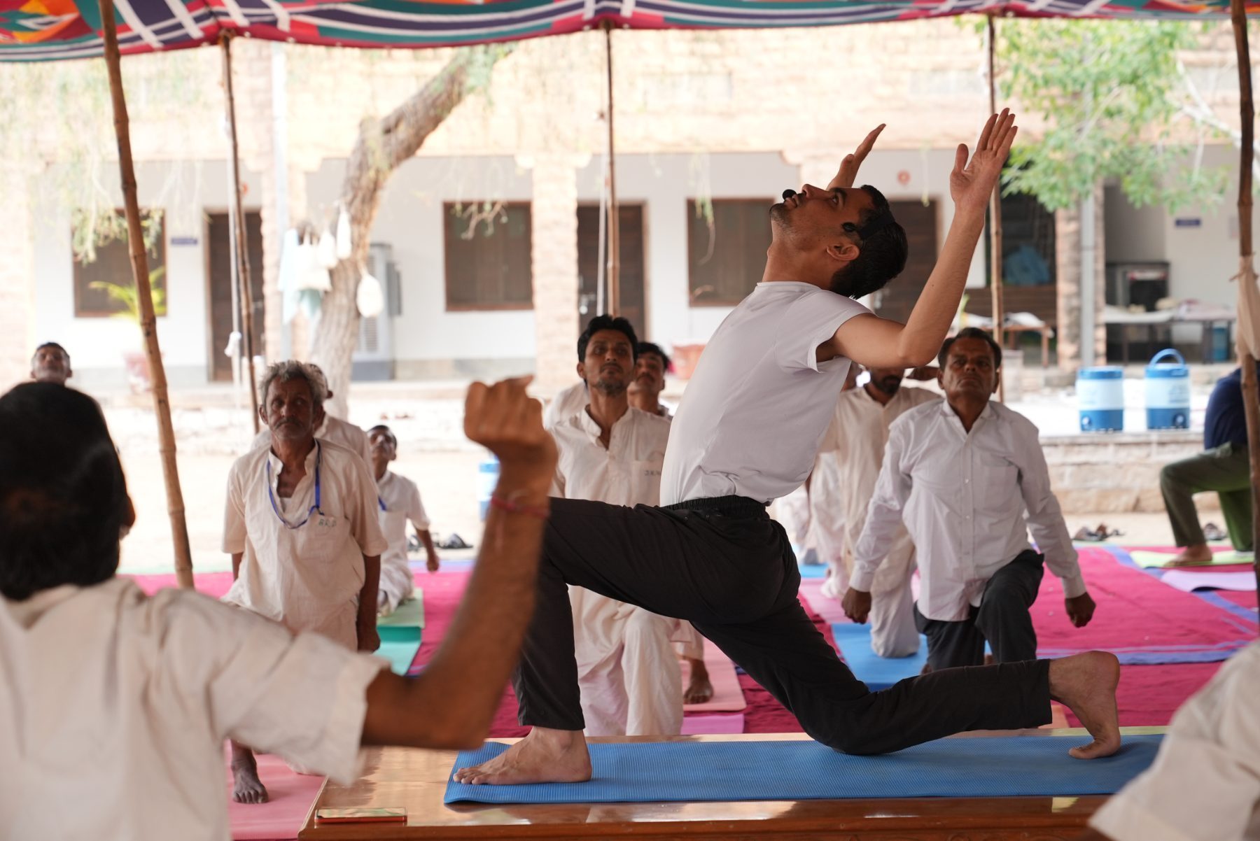 An instructor demonstrating a deep chest-opening lunge on a low wooden platform while the class follows beneath the canopy.