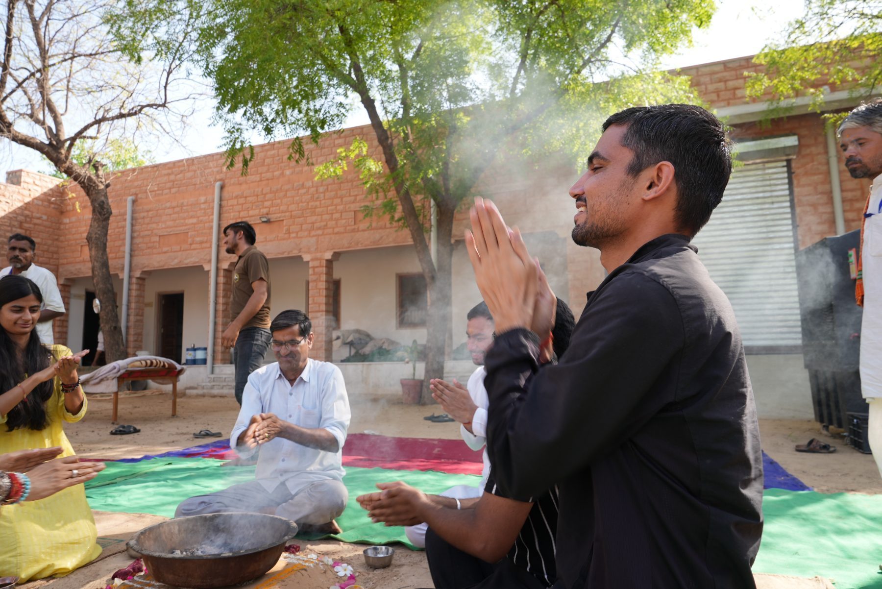 People seated around a small fire with hands raised in prayer, smoke rising between them.