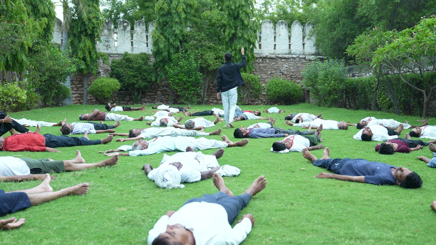 Men lying in shavasana on coloured mats across a lawn, seen from above, instructors standing among them.