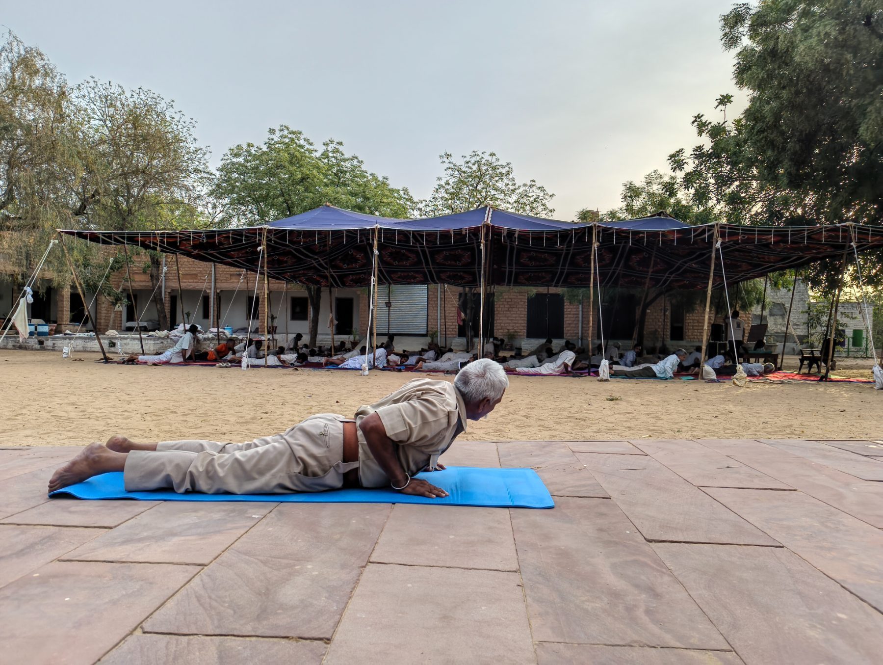 An older man in a khaki uniform holding bhujangasana on a blue mat, with the camp practising under a canopy behind him.