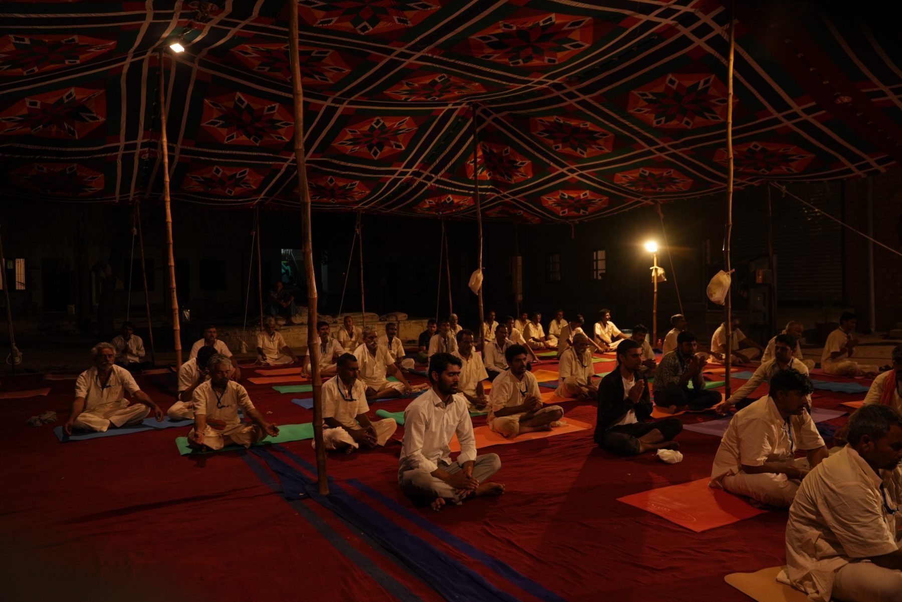 The whole camp seated on coloured mats across a red floor covering at night, lit by a single lamp.