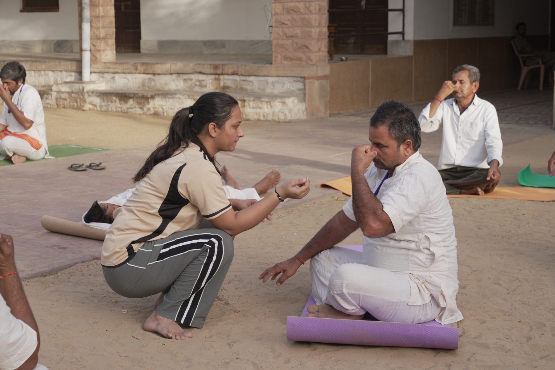 A young woman instructor crouching in a sandy courtyard, showing the hand position for alternate-nostril breathing to an older man seated on a mat.