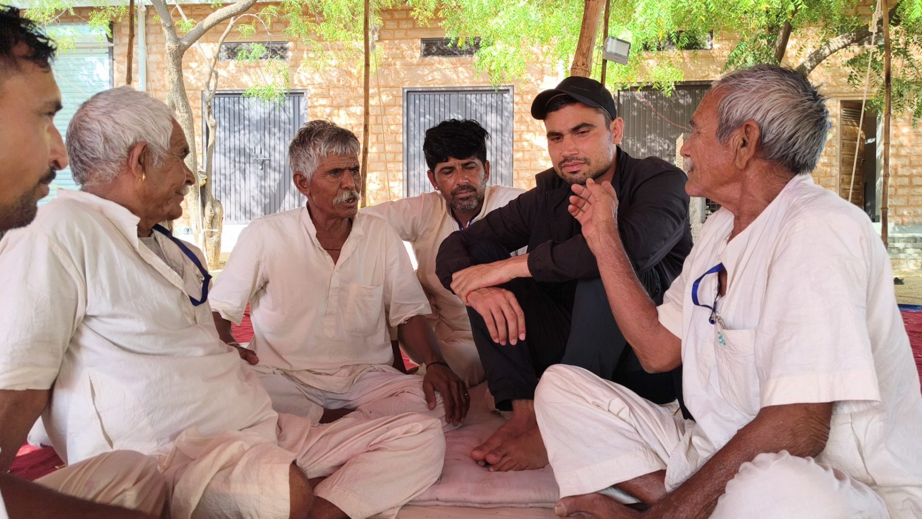 A trainer sitting cross-legged on the ground among a small group of older men, talking with them at eye level.