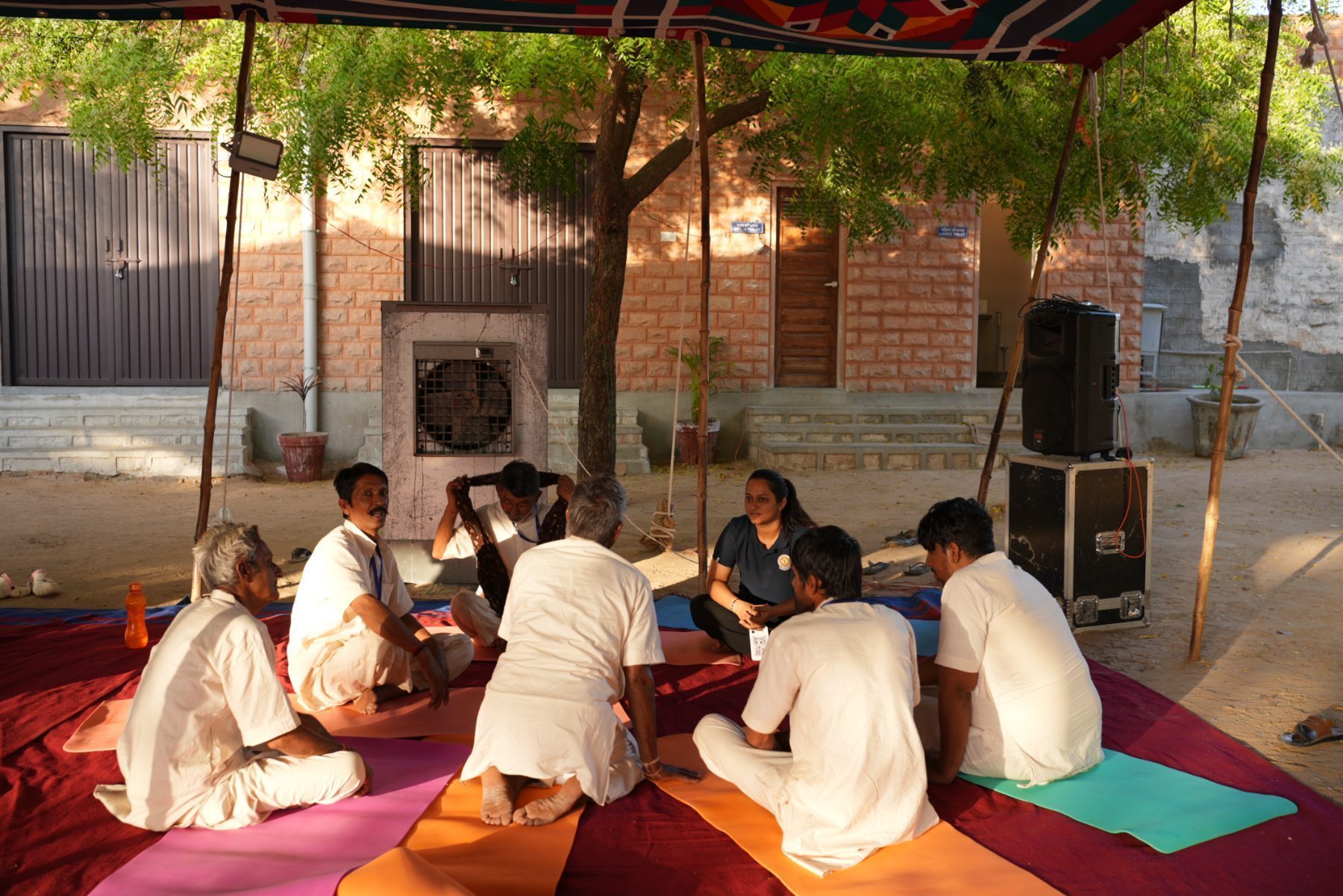 A small circle of participants seated on coloured mats under the canopy with an instructor sitting among them, one man practising with a cloth at his face.