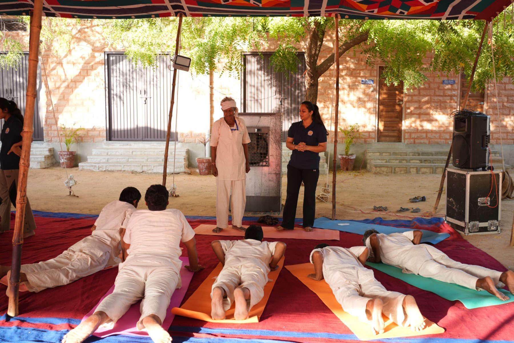 Participants in bhujangasana on coloured mats under the canopy, an older man leading them, instructors standing close by.