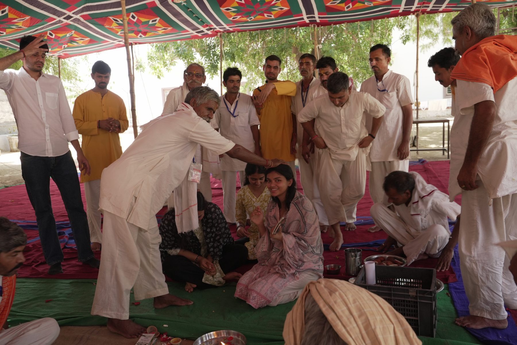 Under the canopy, an older participant leaning down to place his hand on the head of a young woman from the team, who is smiling, with a lamp and offering plate on the ground.
