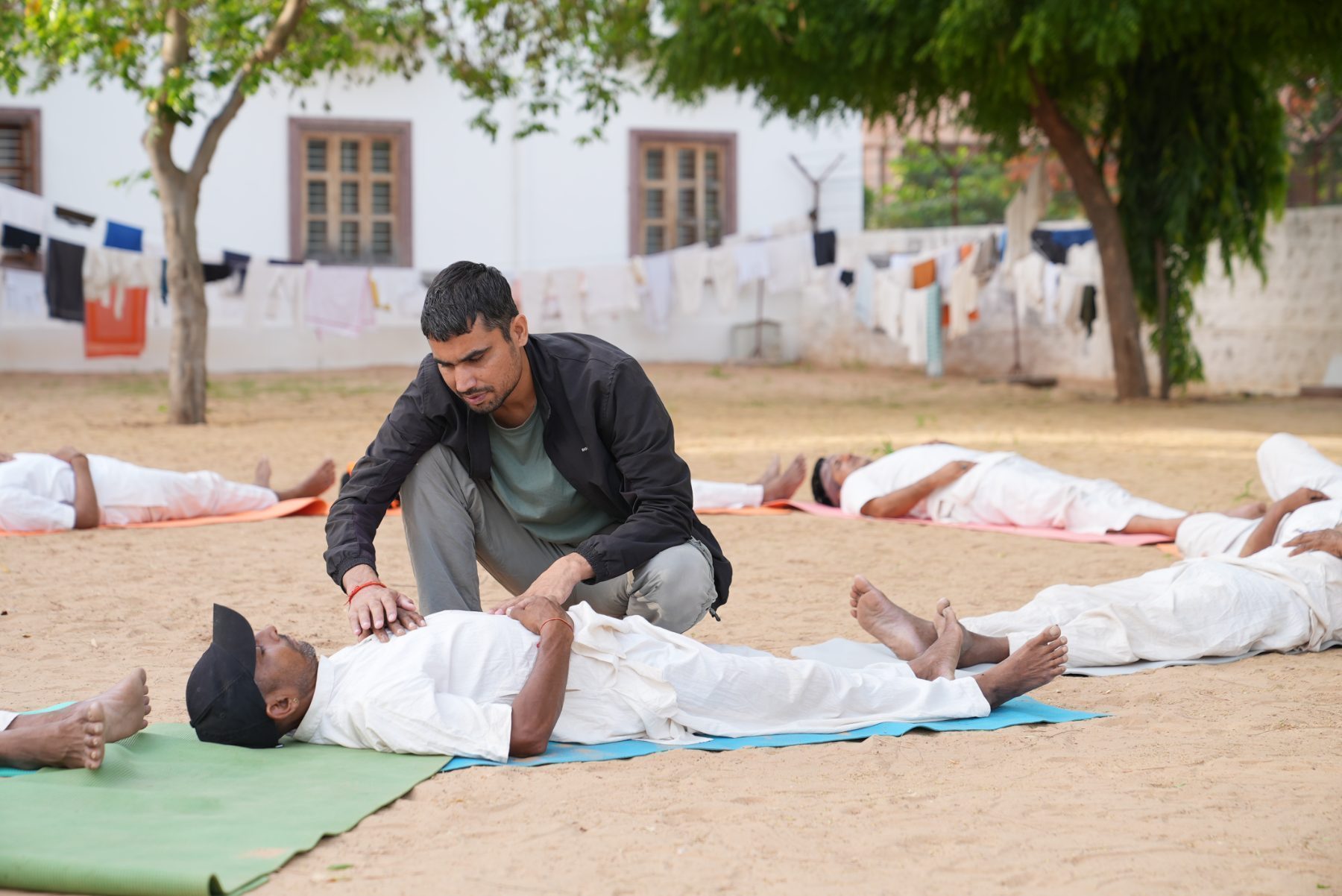 An instructor kneeling in the sand with both hands resting on the chest of a man lying in shavasana wearing a black cap, others resting nearby.