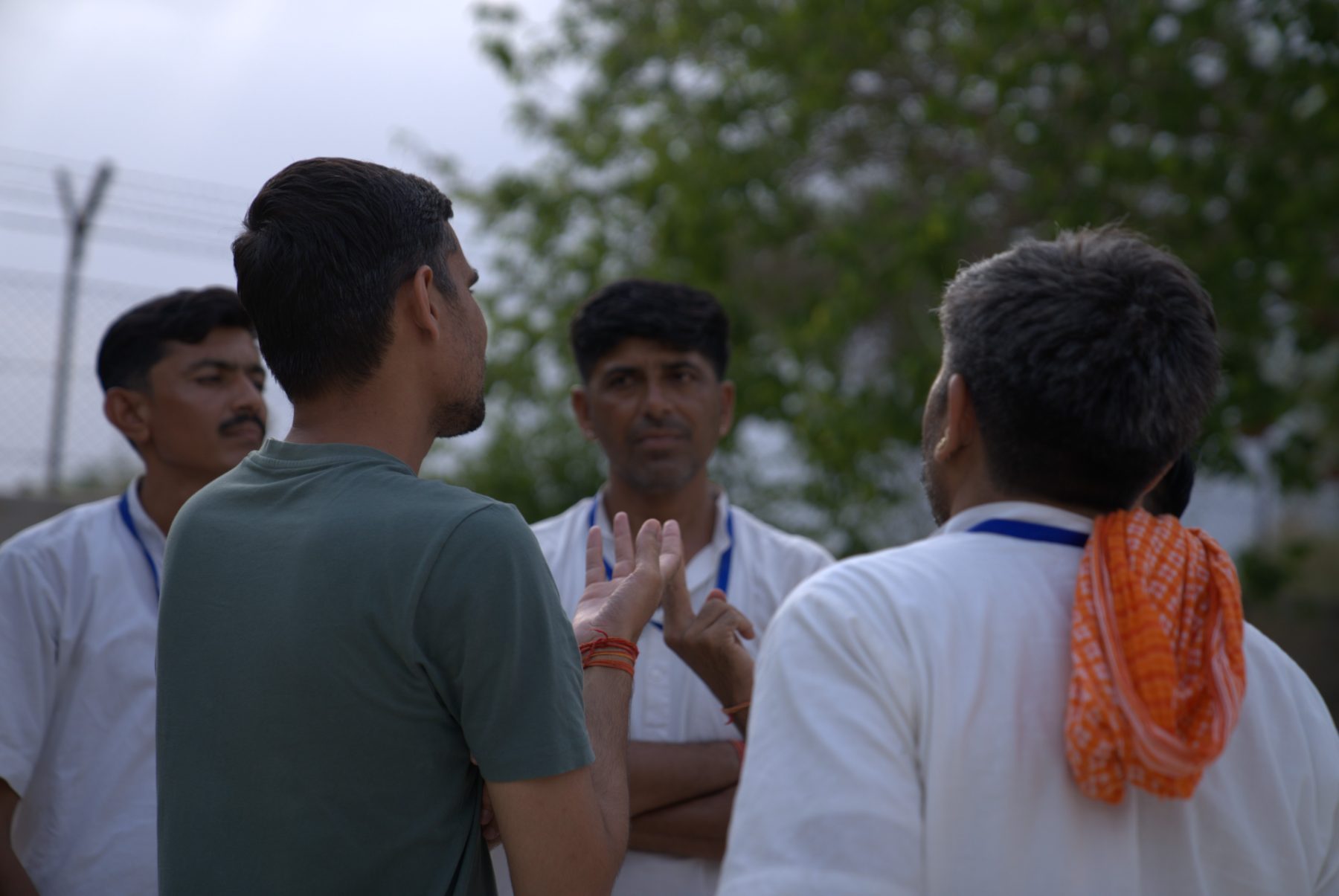 Under a grey sky, a member of the team standing with a small group of participants, one of them speaking with his hand raised, faces serious.