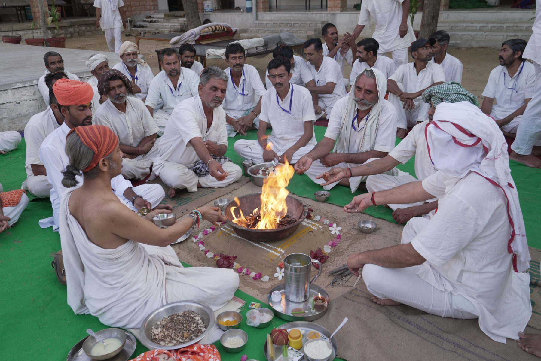 A havan in progress: a copper vessel with fire on a bed of sand ringed with flower petals, offerings being made by seated participants.