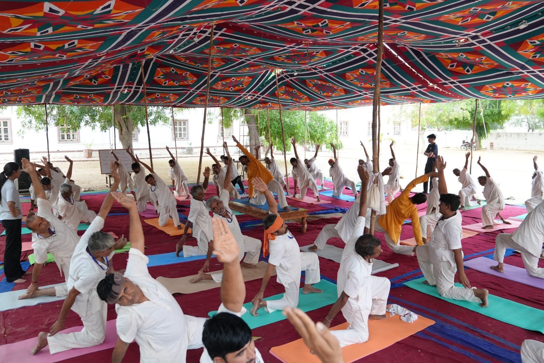 A full class holding a demanding kneeling posture with arms raised beneath a bright canopy, an instructor demonstrating on a low wooden bench.