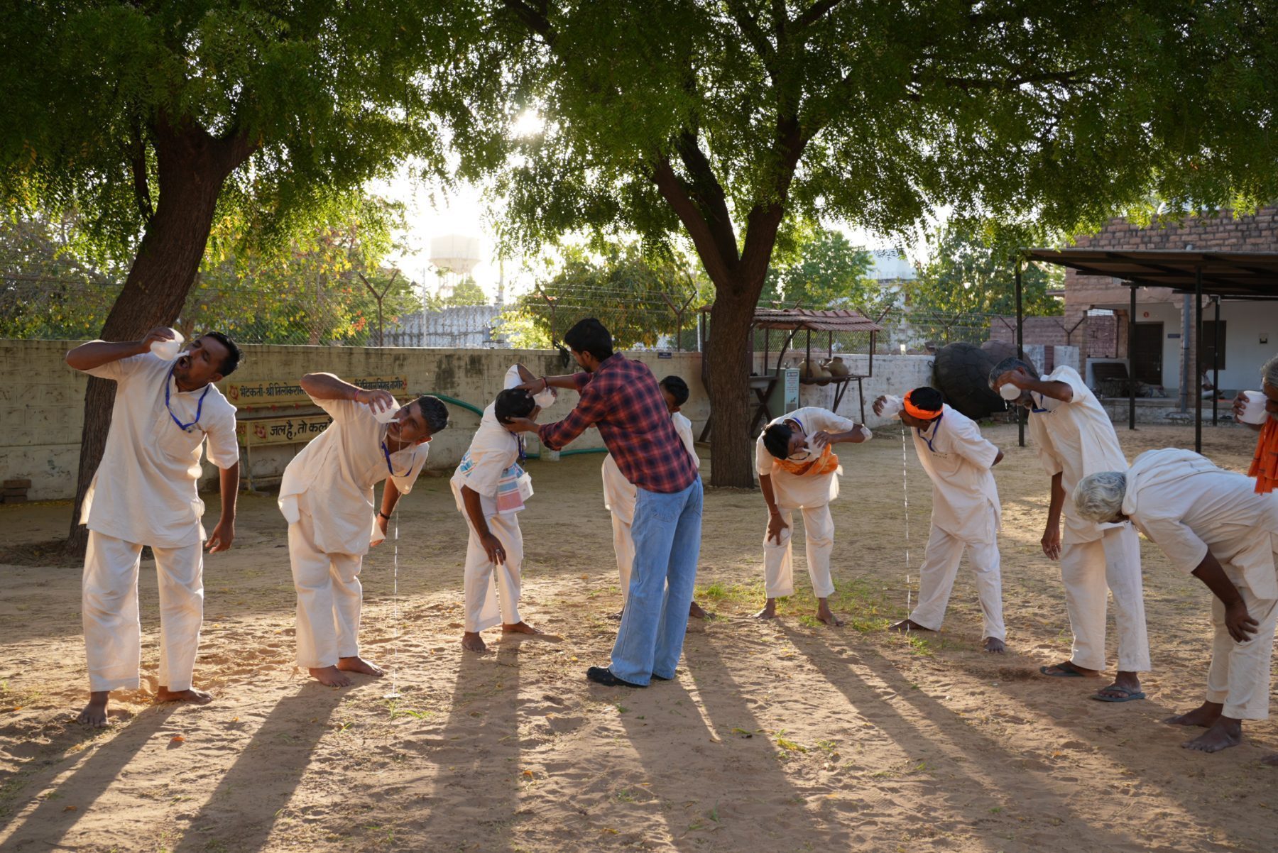 Men bent forward in a sandy yard at sunrise during a cleansing practice, one of the team pouring water and steadying a head, long shadows across the ground.
