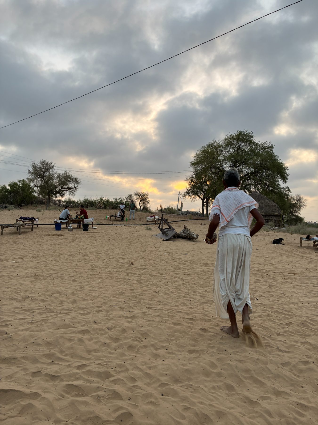 An elder in white walking away across open sand at dusk toward a distant group under a heavy sky, with a single break of gold behind the trees.