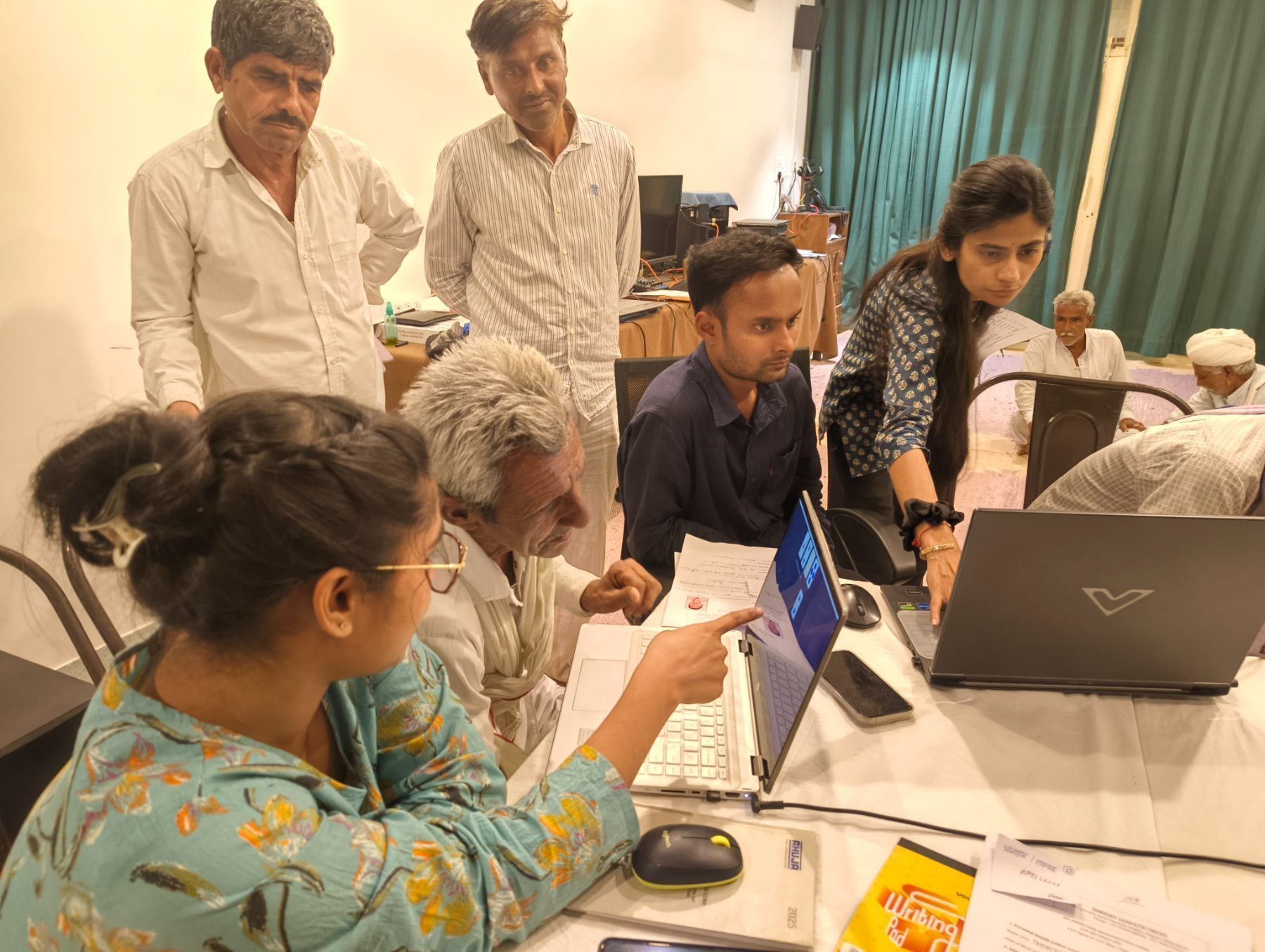 Two young women from the research team guiding participants through a computer-based assessment, an older man concentrating at the screen, others waiting their turn behind.
