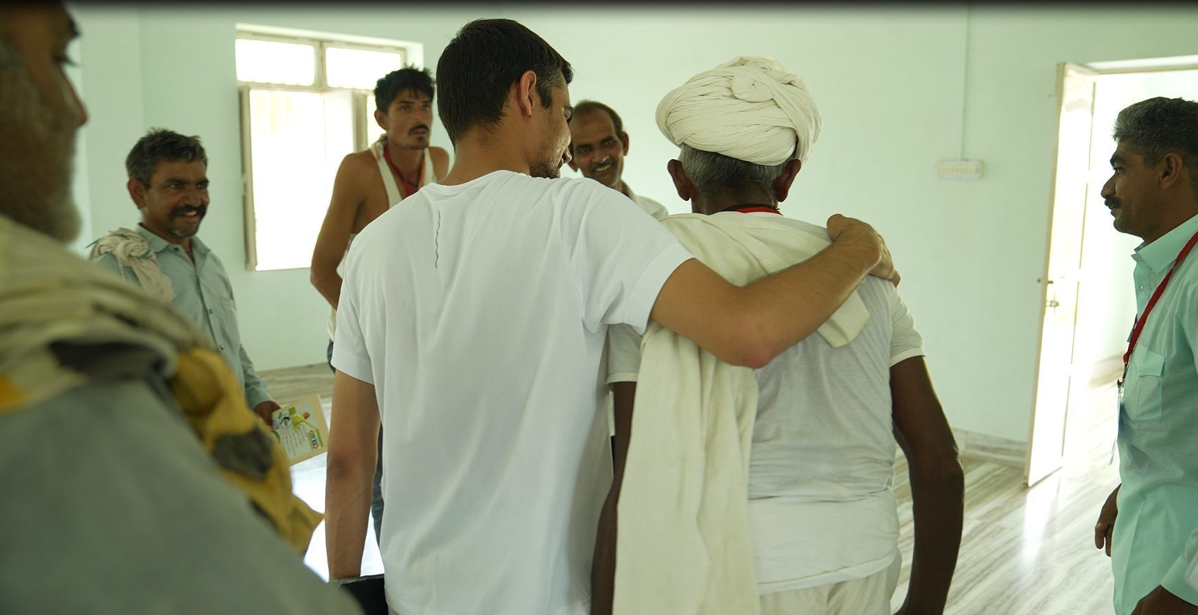 Inside a bright room, a younger man walks out with his arm around the shoulders of an older man in a white turban. Others nearby are smiling.