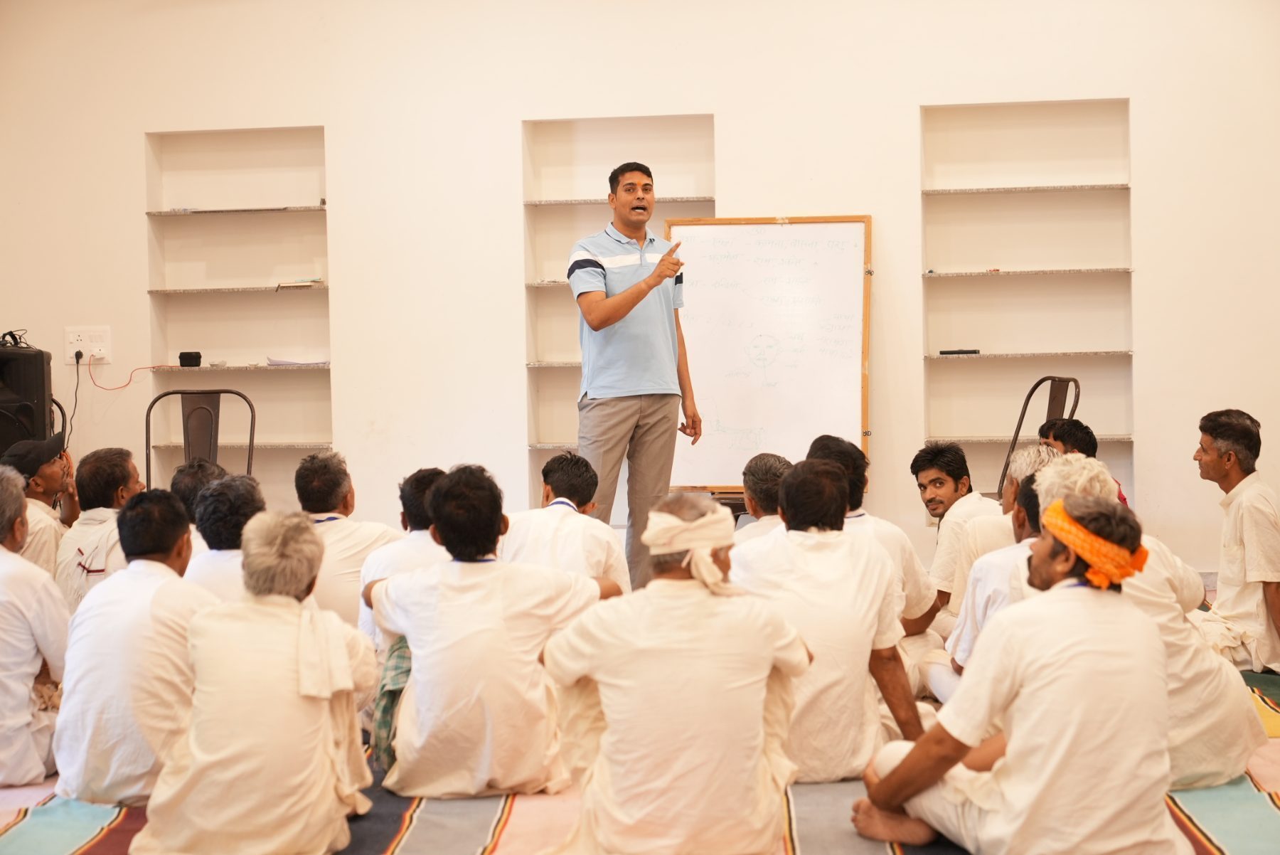 A teacher at a whiteboard covered in Hindi notes, explaining to men seated on the floor.